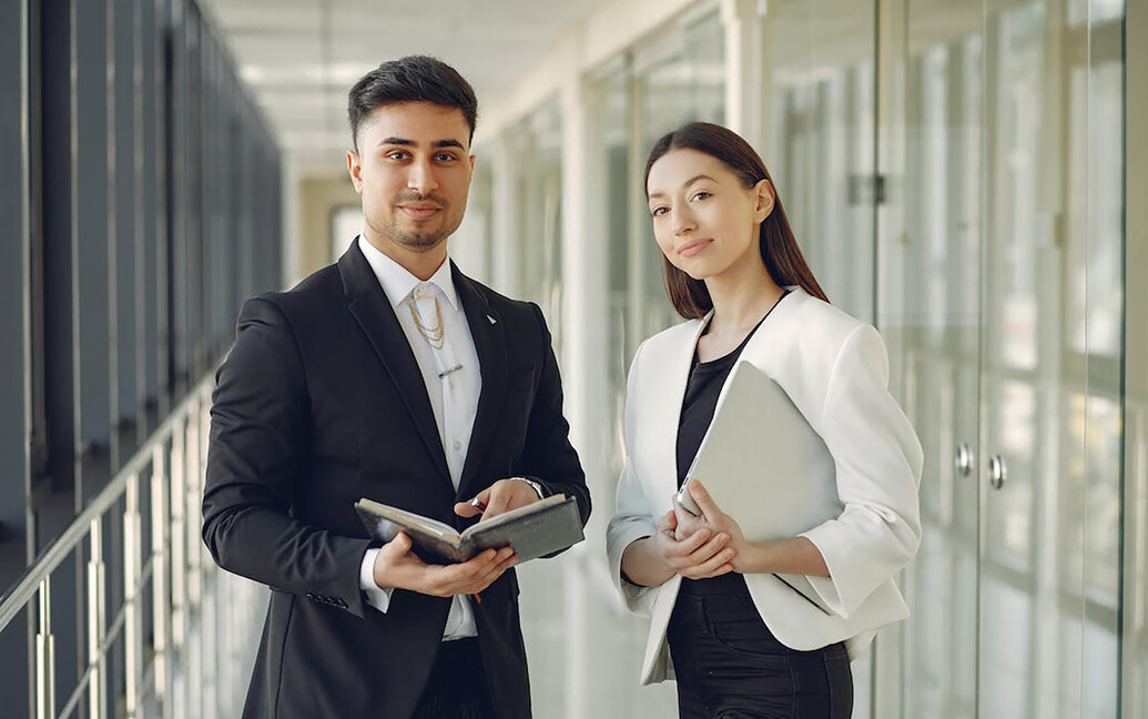 Business psychology co-workers a smiling male and a female both wearing suits hold books in an office corridor