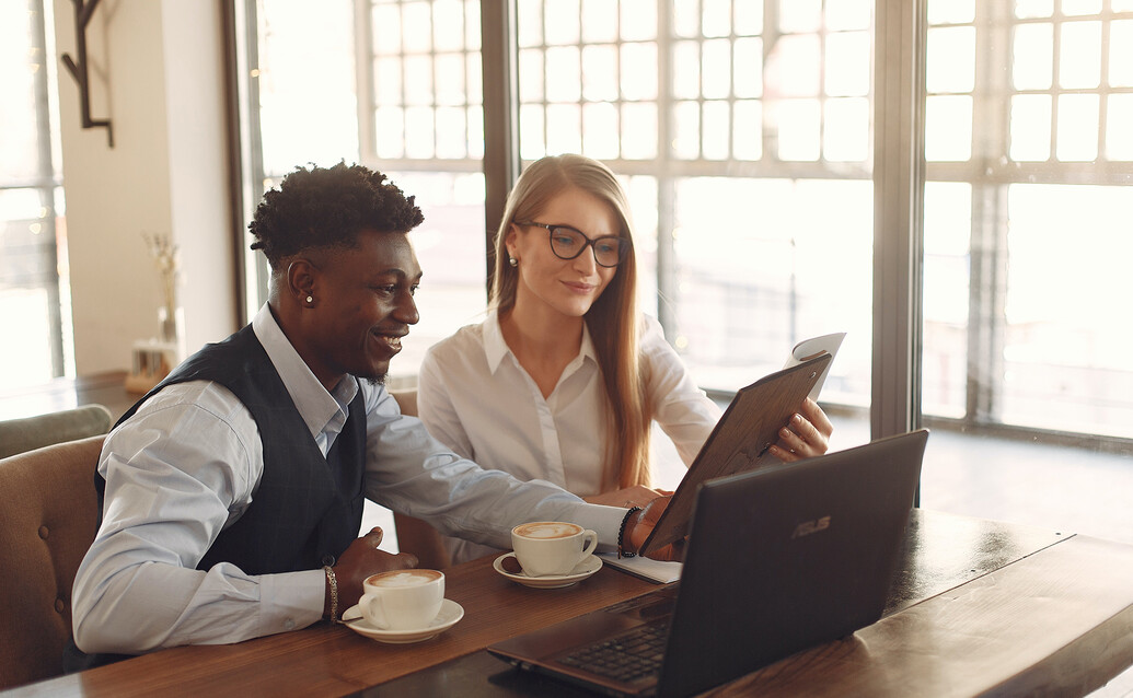 Occupational and organisational psychology A smiling male and female sitting at a table in front of a laptop and two coffee cups looking at a notebook